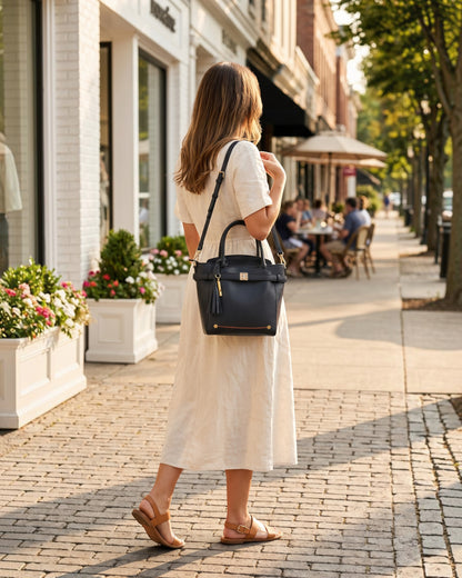 Woman wearing a black Montemaré Mayfair Tall Crossbody bag with a tassel, walking on a sunny city sidewalk.