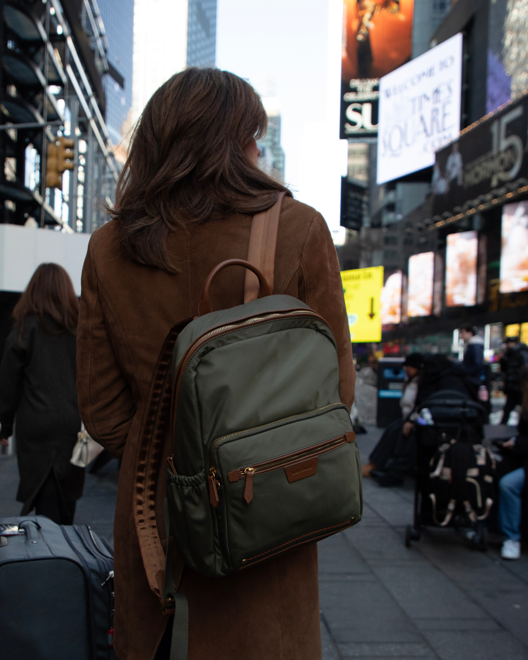 Person with a green backpack walking in a busy city street with advertisements and people around.
