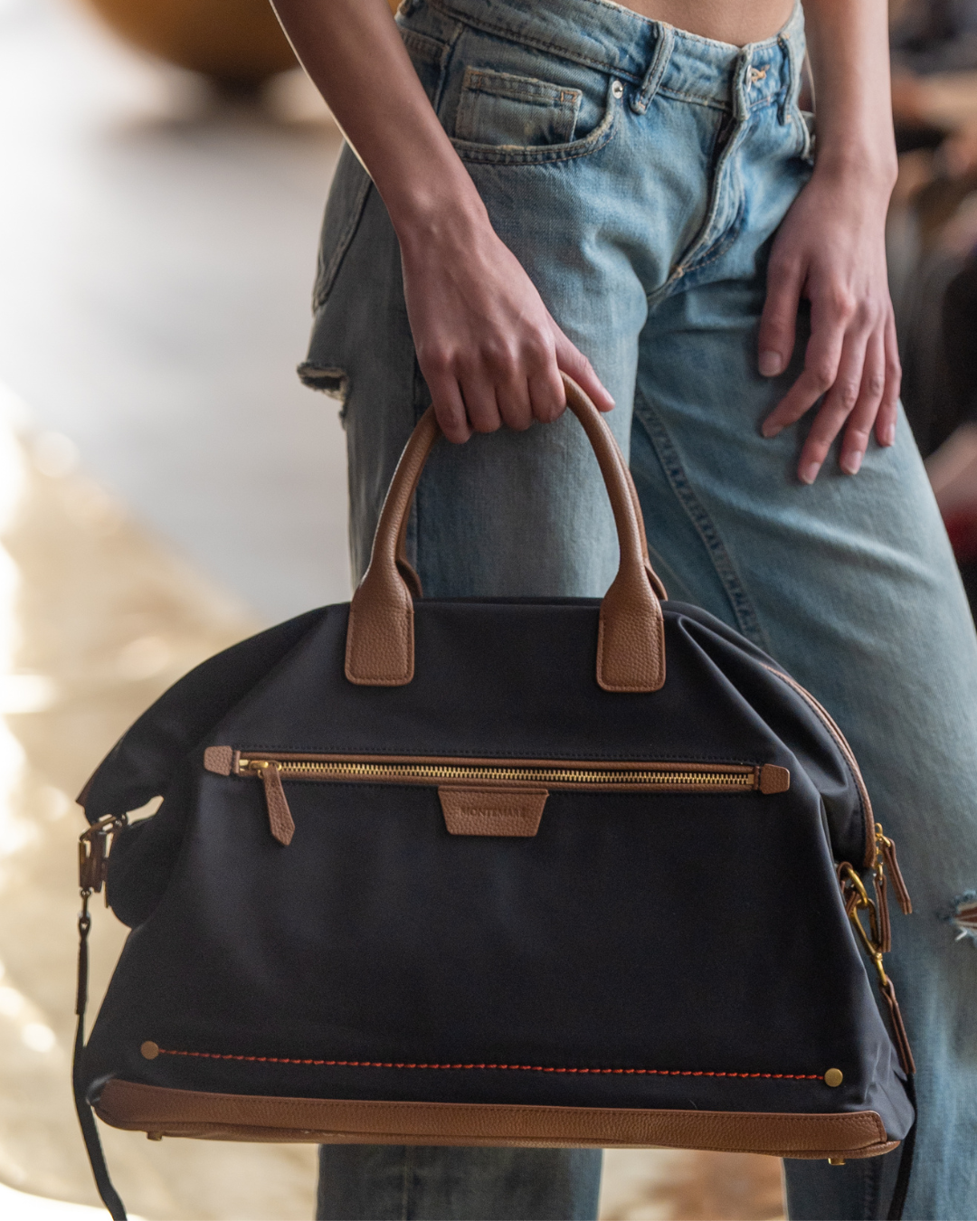 Person holding a black handbag with brown handles and trim, wearing blue jeans.