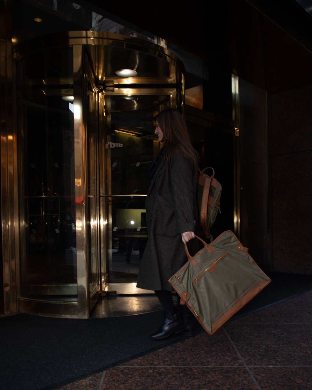 Woman carrying a green olive garment bag entering a revolving door in a dimly lit setting