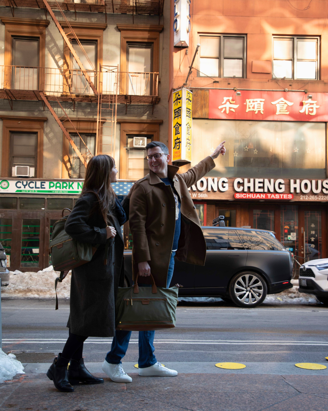 Two people with travel bags on a city street with a building and sign in the background.