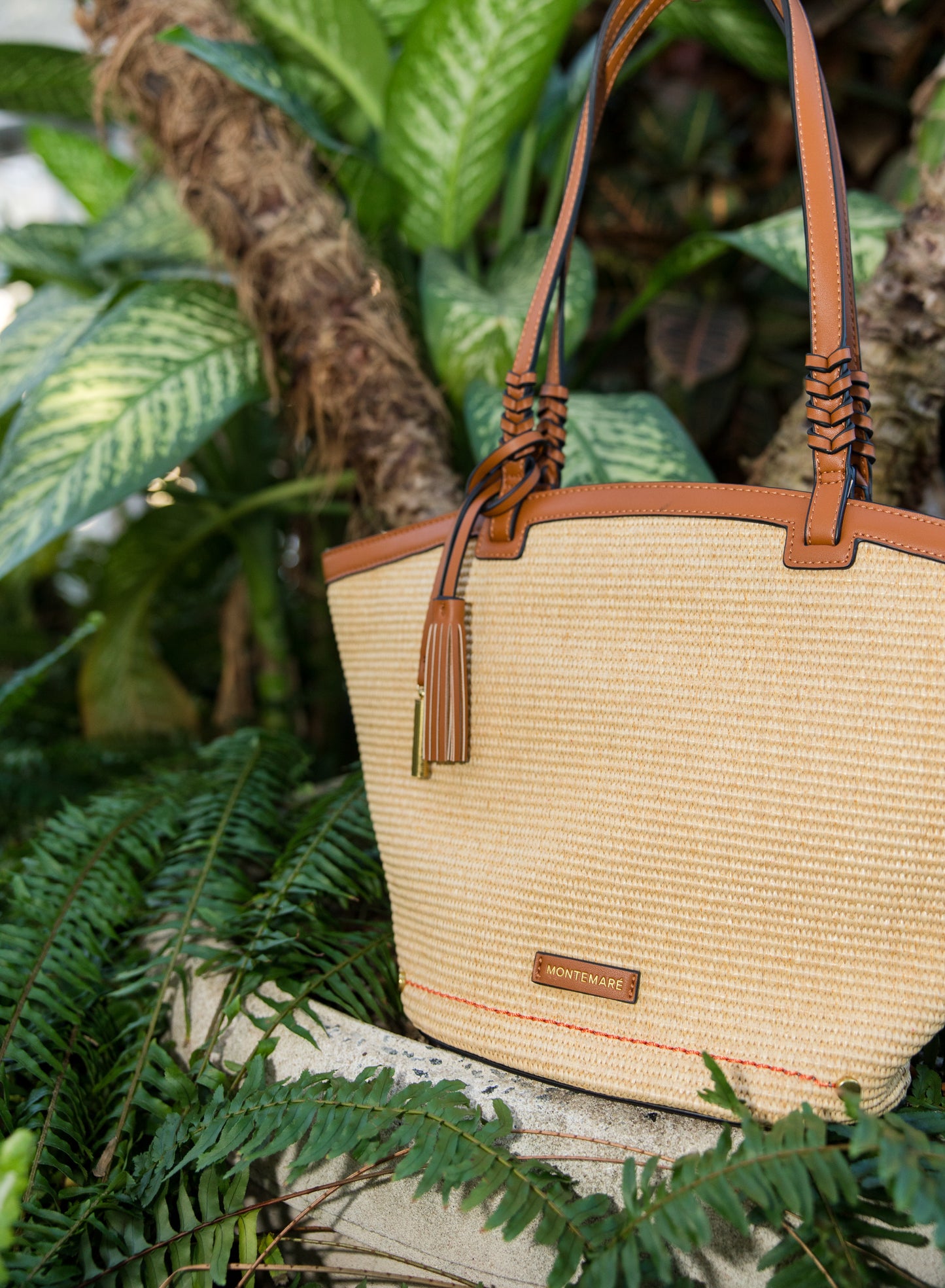 Beige straw handbag with brown leather straps and accents, placed on a stone surface with green foliage in the background.