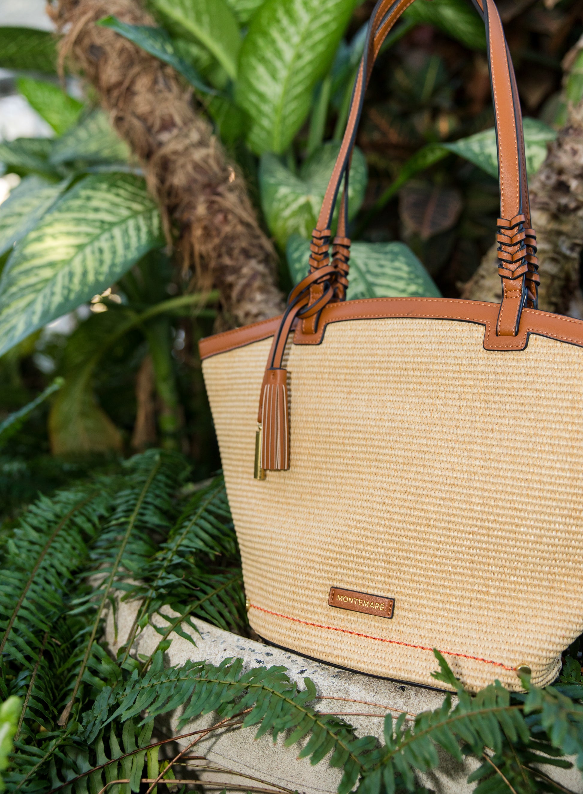 Beige straw handbag with brown leather straps and accents, placed on a stone surface with green foliage in the background.