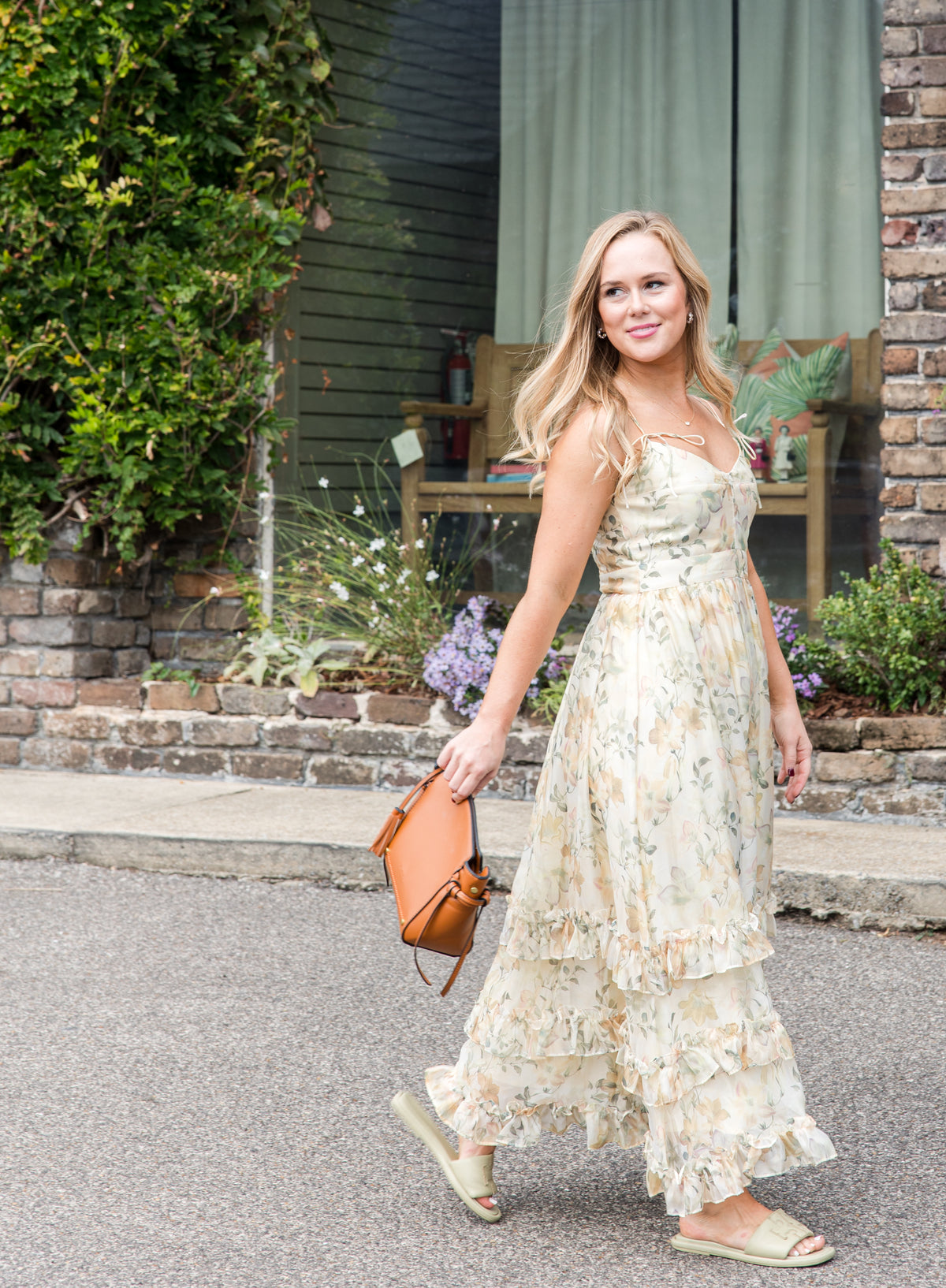 Woman in a floral dress with brown handbag walking outdoors with a brick building and greenery in the background