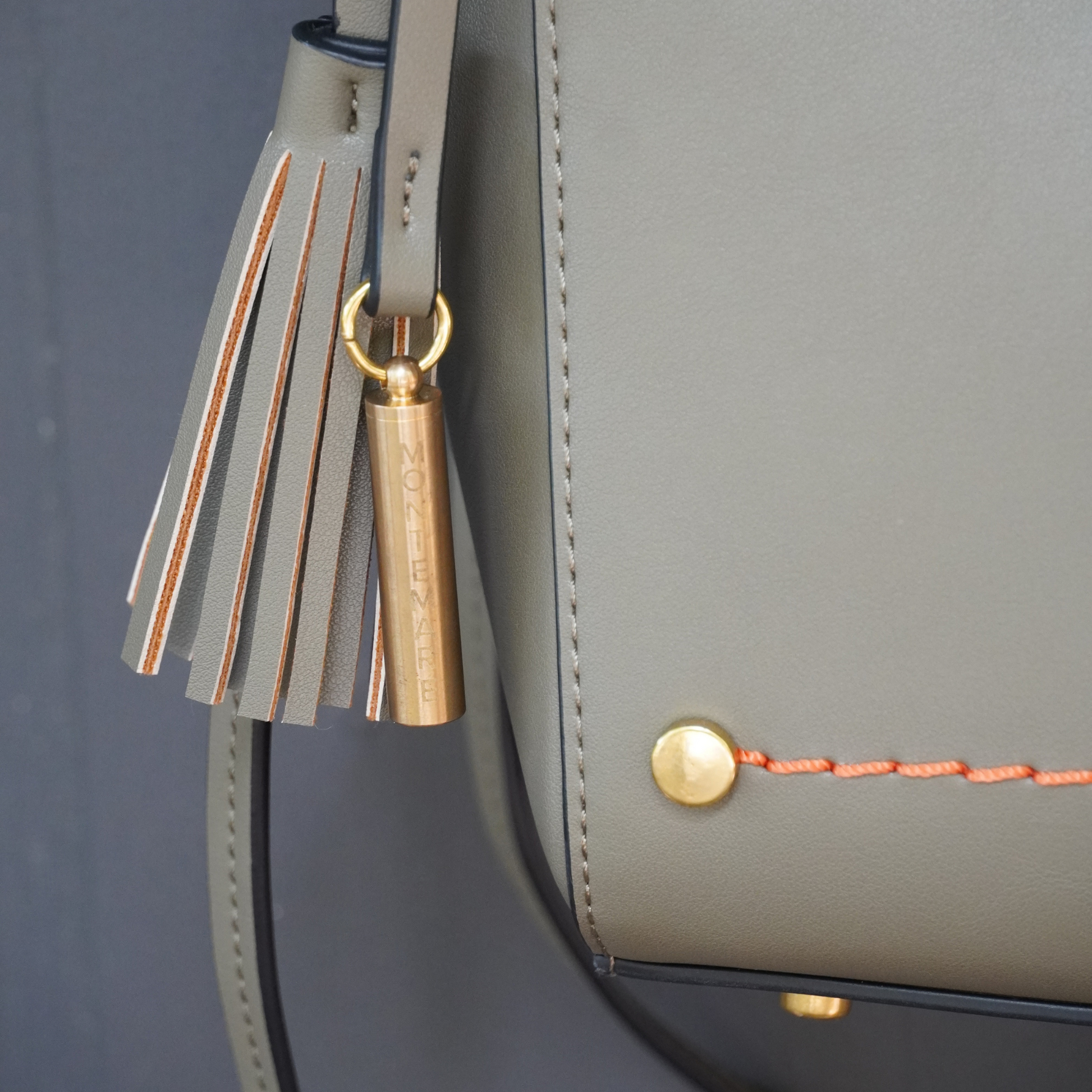 Close up shot of Montemare Roma Crossbody in olive colorway with orange thread accent, whistle tassel on a gray background