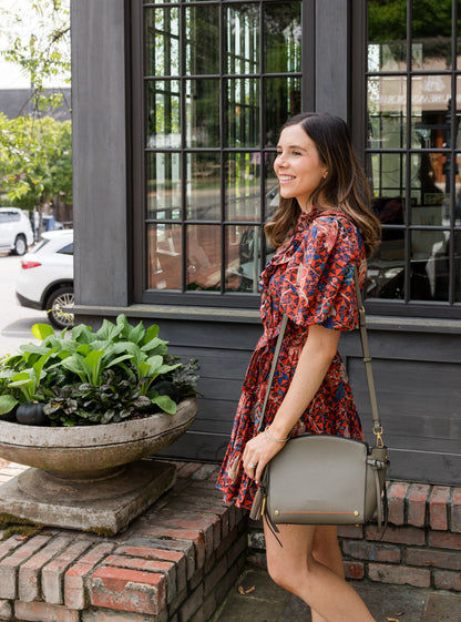 Woman in a floral dress with olive green handbag standing outside a building with large windows.