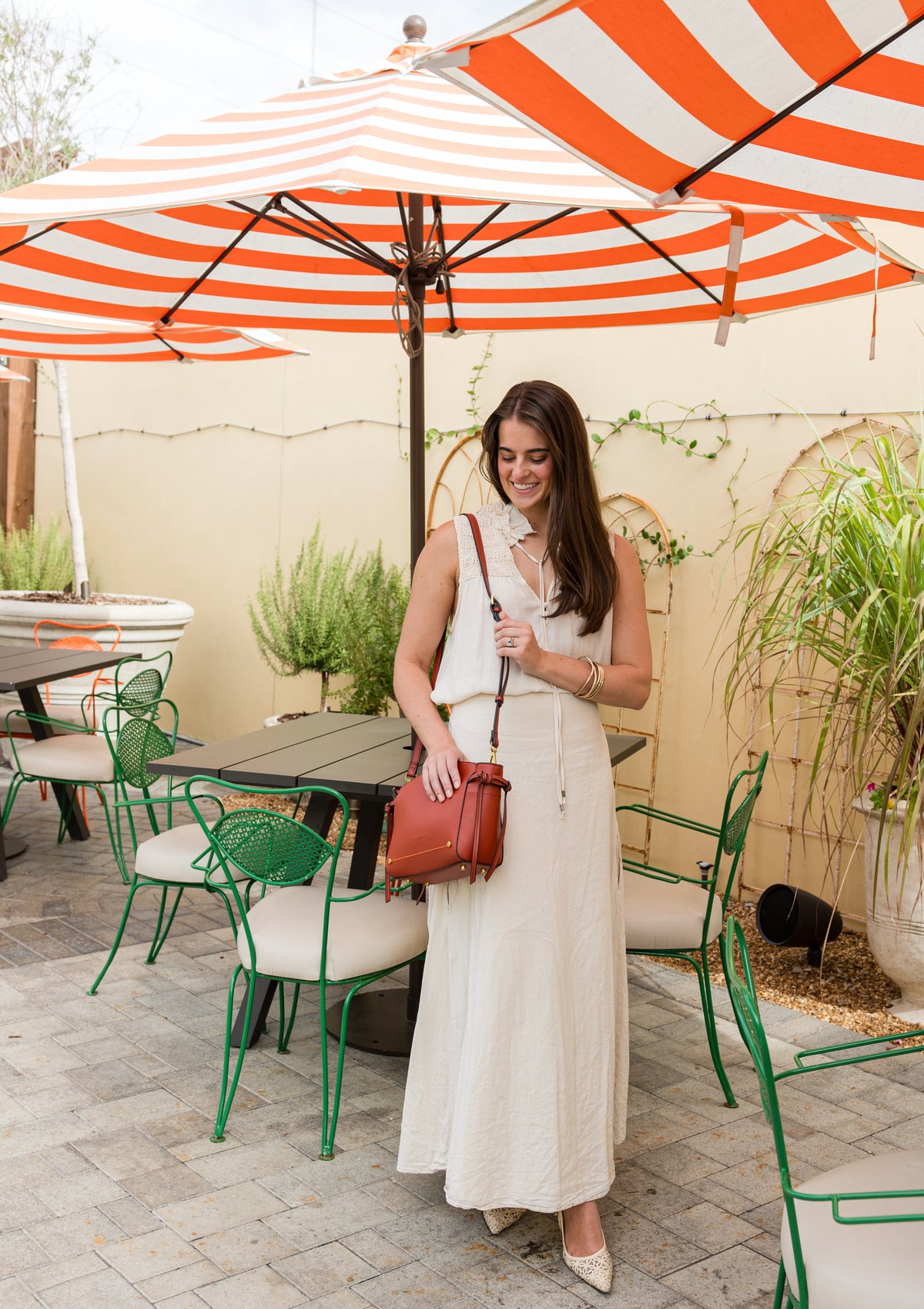 Woman in a white dress with a terracotta handbag standing under a red and white striped umbrella in an outdoor setting.