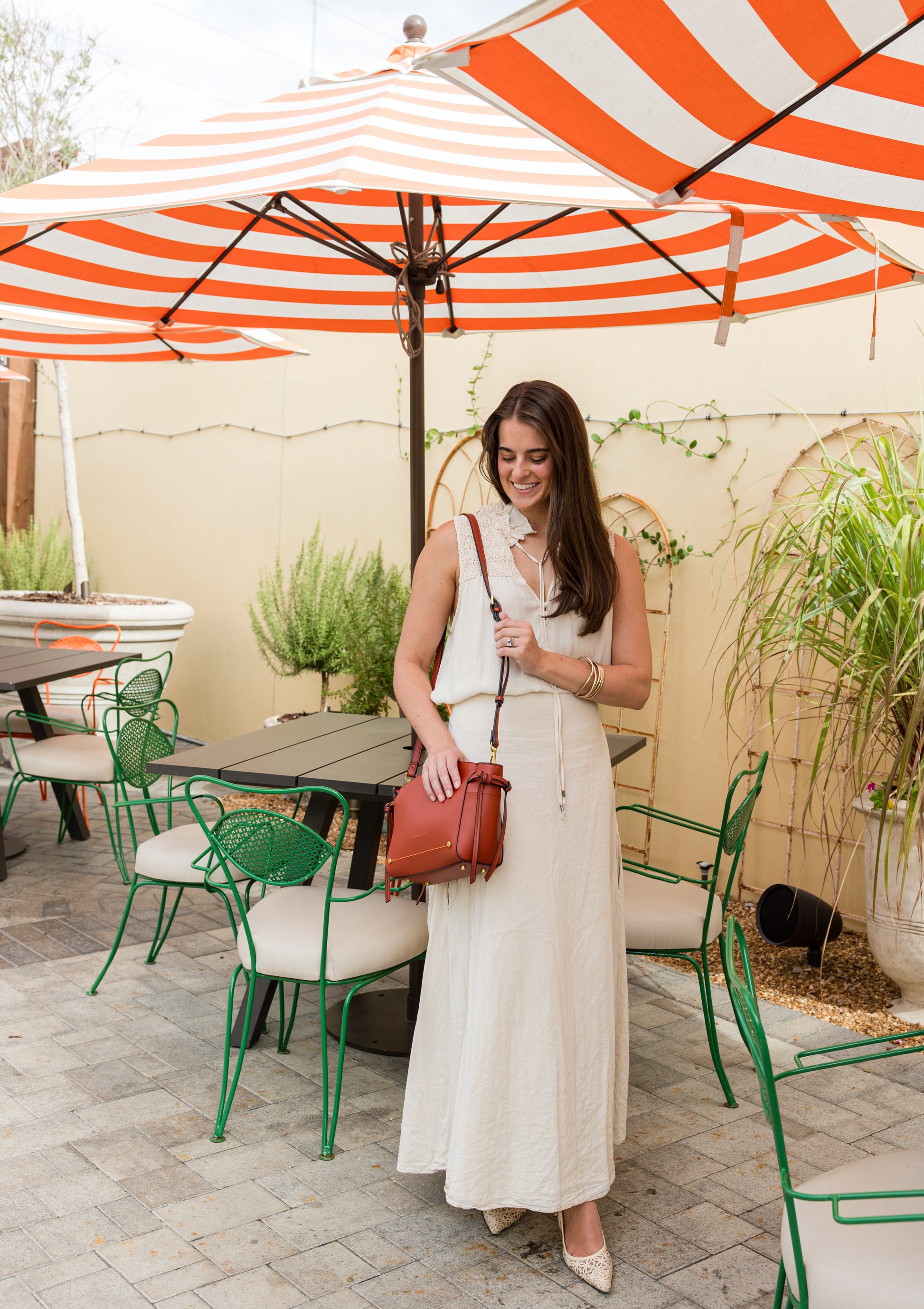 Woman in a white dress with a terracotta handbag standing under a red and white striped umbrella in an outdoor setting.