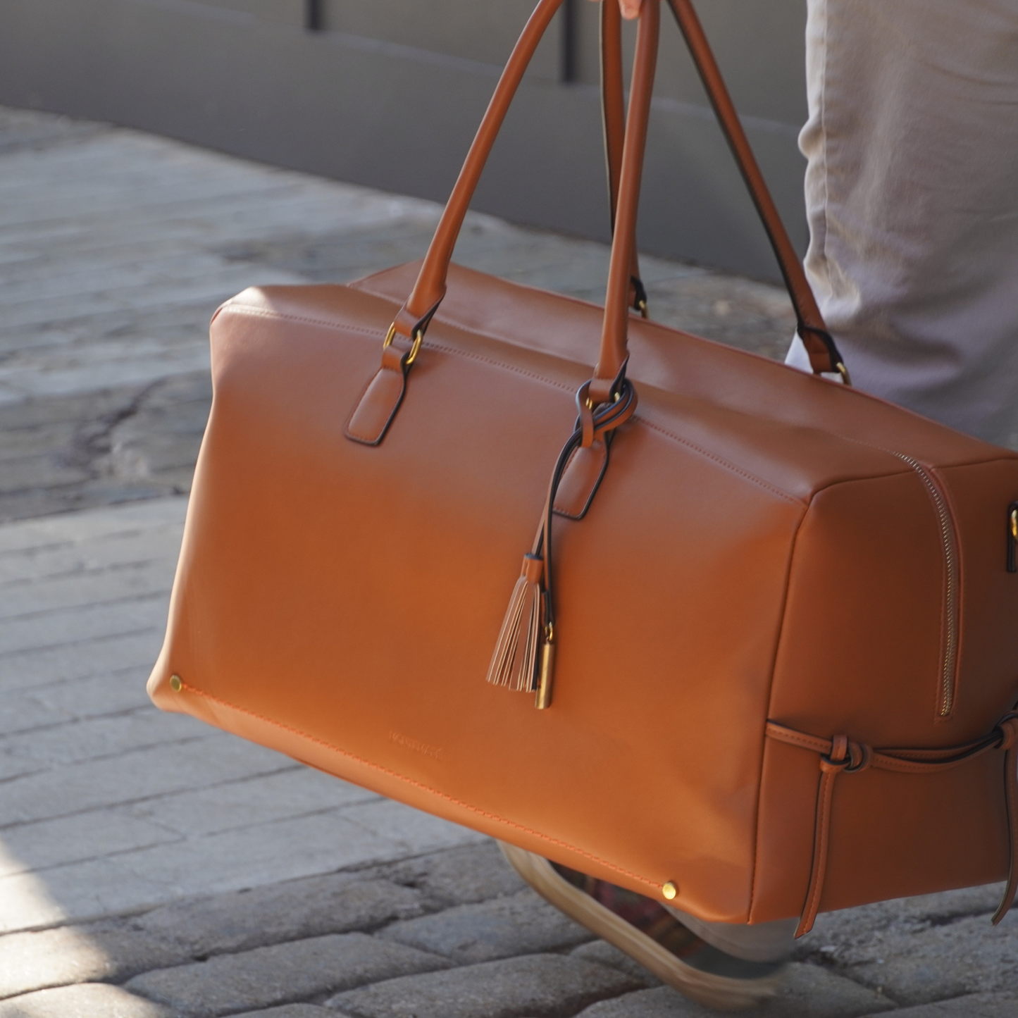 A brown vegan leather duffel bag with a trolley sleeve and a crossbody strap being wheeled on a cobblestone street.