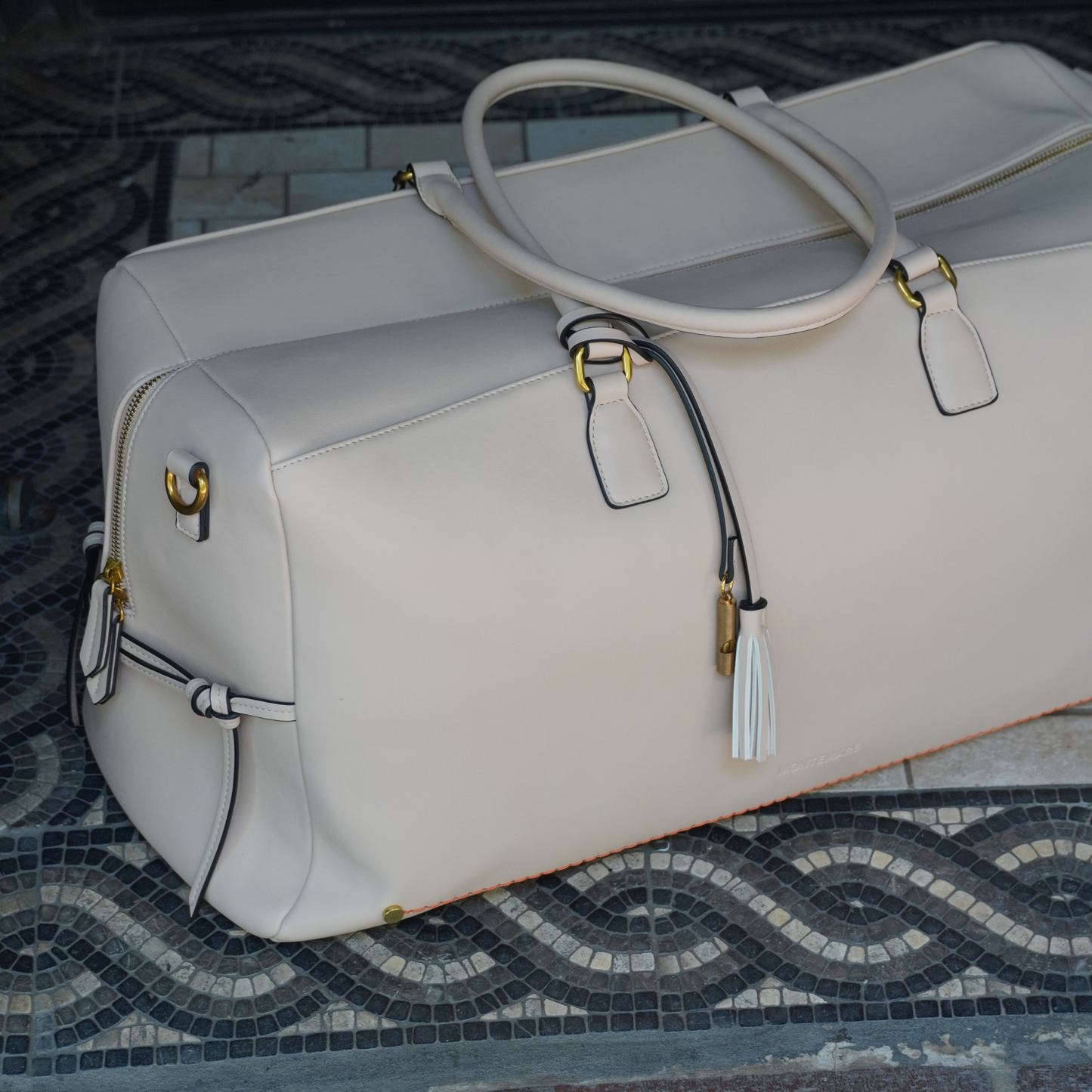 An ivory colored vegan leather duffel bag with a trolley sleeve and a crossbody strap being wheeled on a cobblestone street.