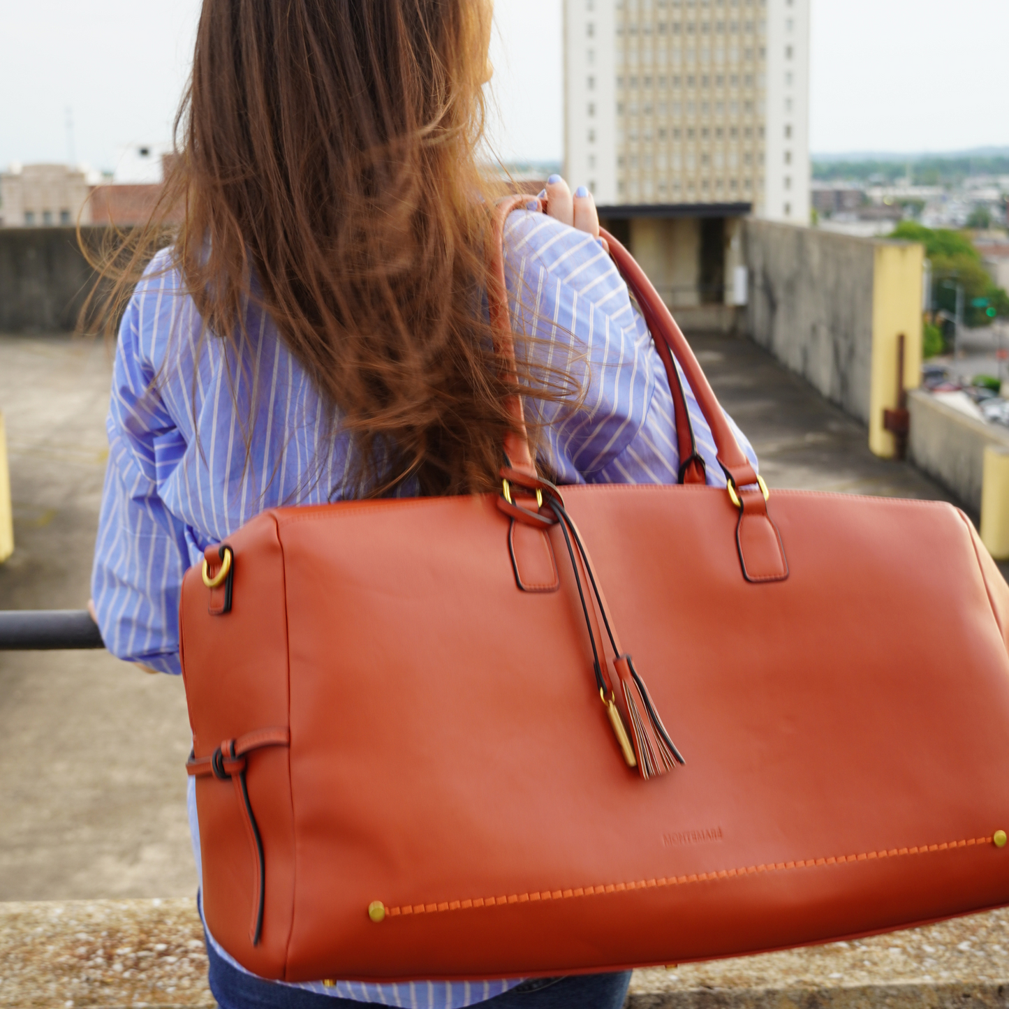A woman wearing blue striped sleeves holding a Montemare Duffel Bag in terracotta colorway on a building rooftop