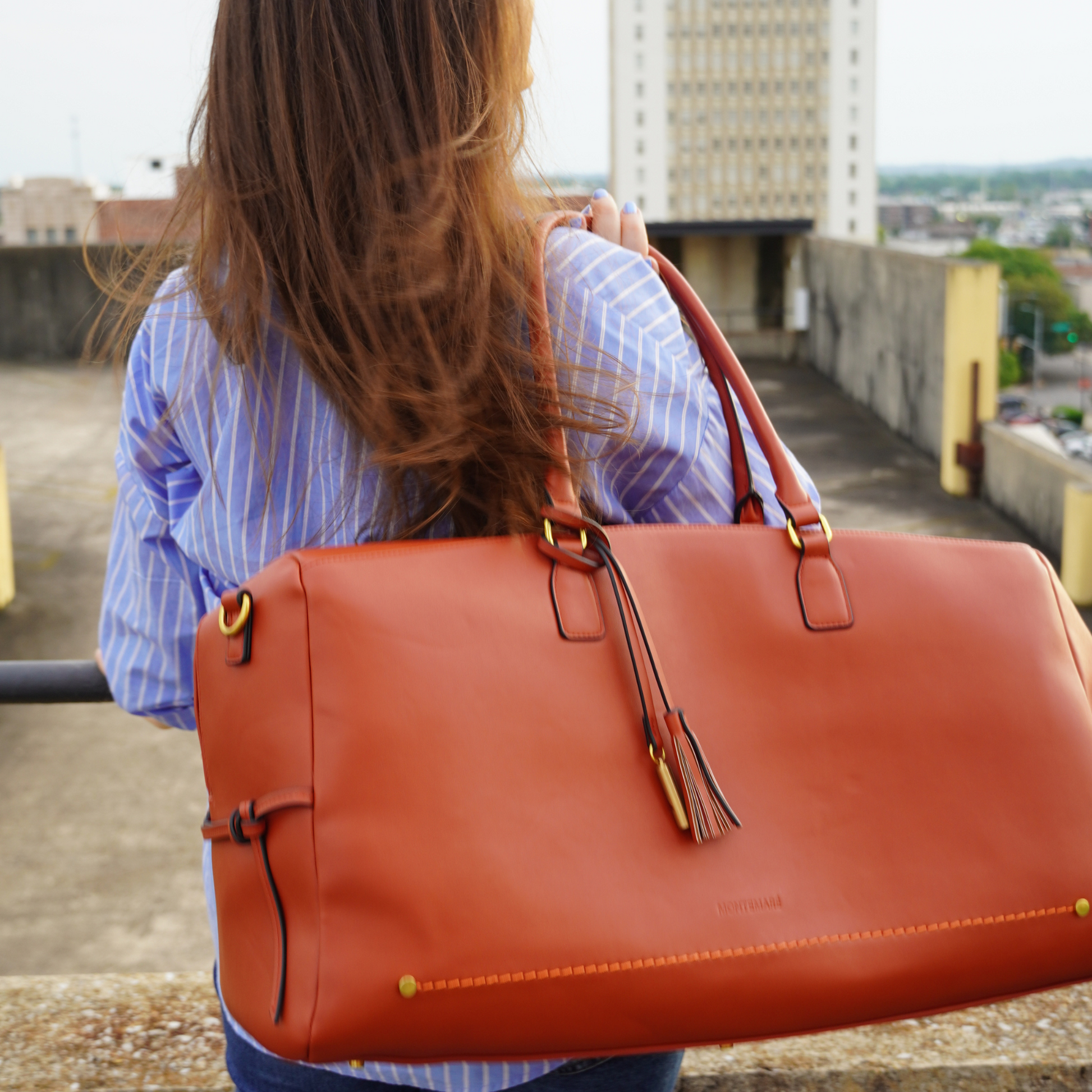 A woman wearing blue striped sleeves holding a Montemare Duffel Bag in terracotta colorway on a building rooftop