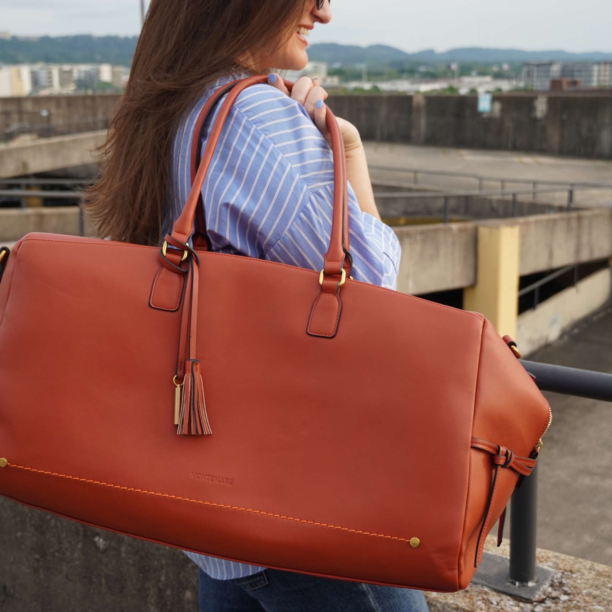 A brown vegan leather duffel bag with a trolley sleeve and a crossbody strap being wheeled on a parking building rooftop.