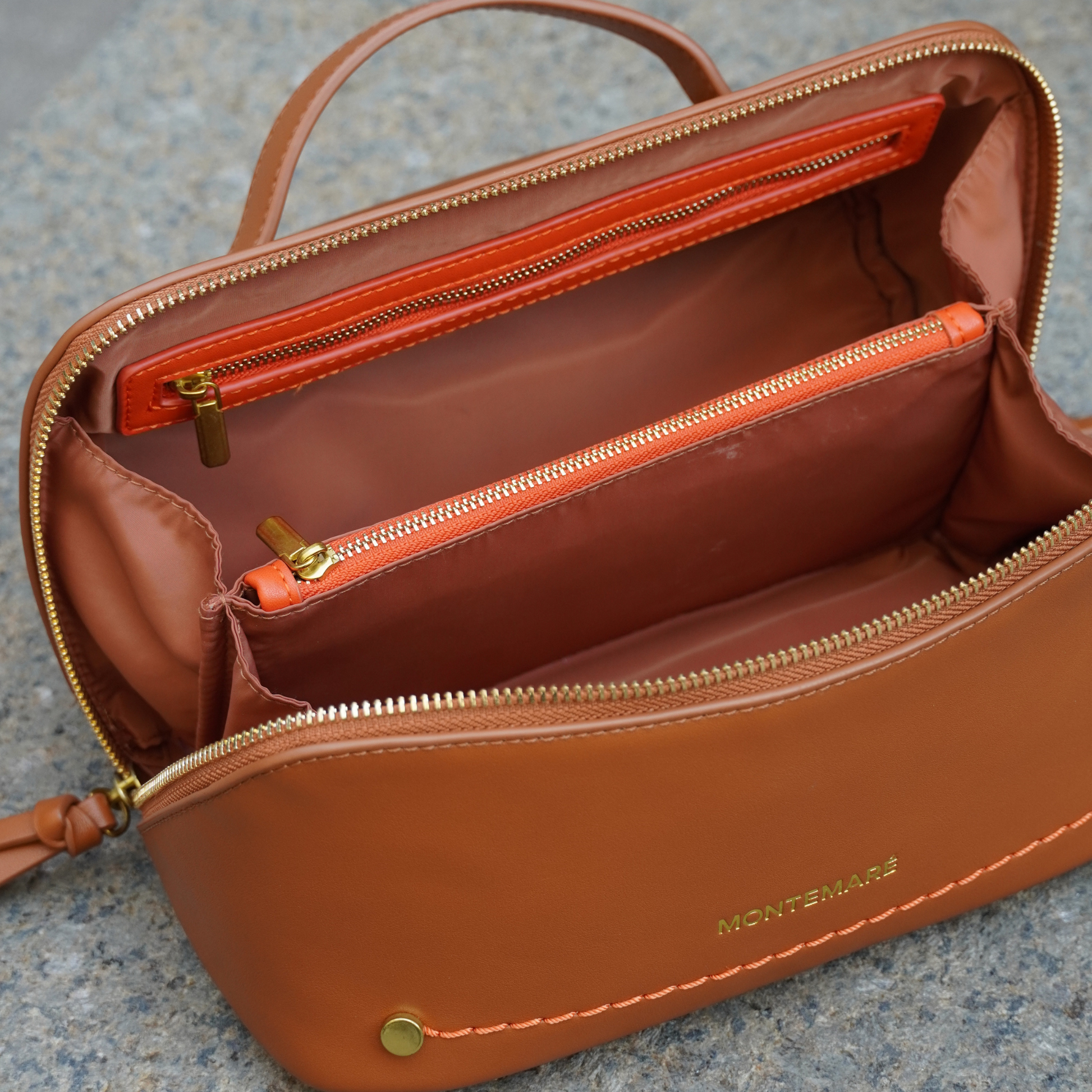 A cognac Roma Foldout Toiletry Case with vegan leather details and orange zippers, displayed on a marble surface with plants in the background.