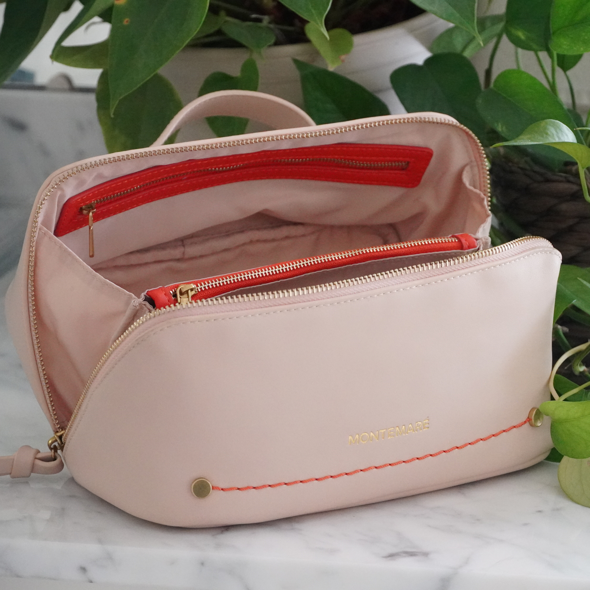 An ivory Roma Foldout Toiletry Case with vegan leather details and orange zippers, displayed on a marble surface with plants in the background.