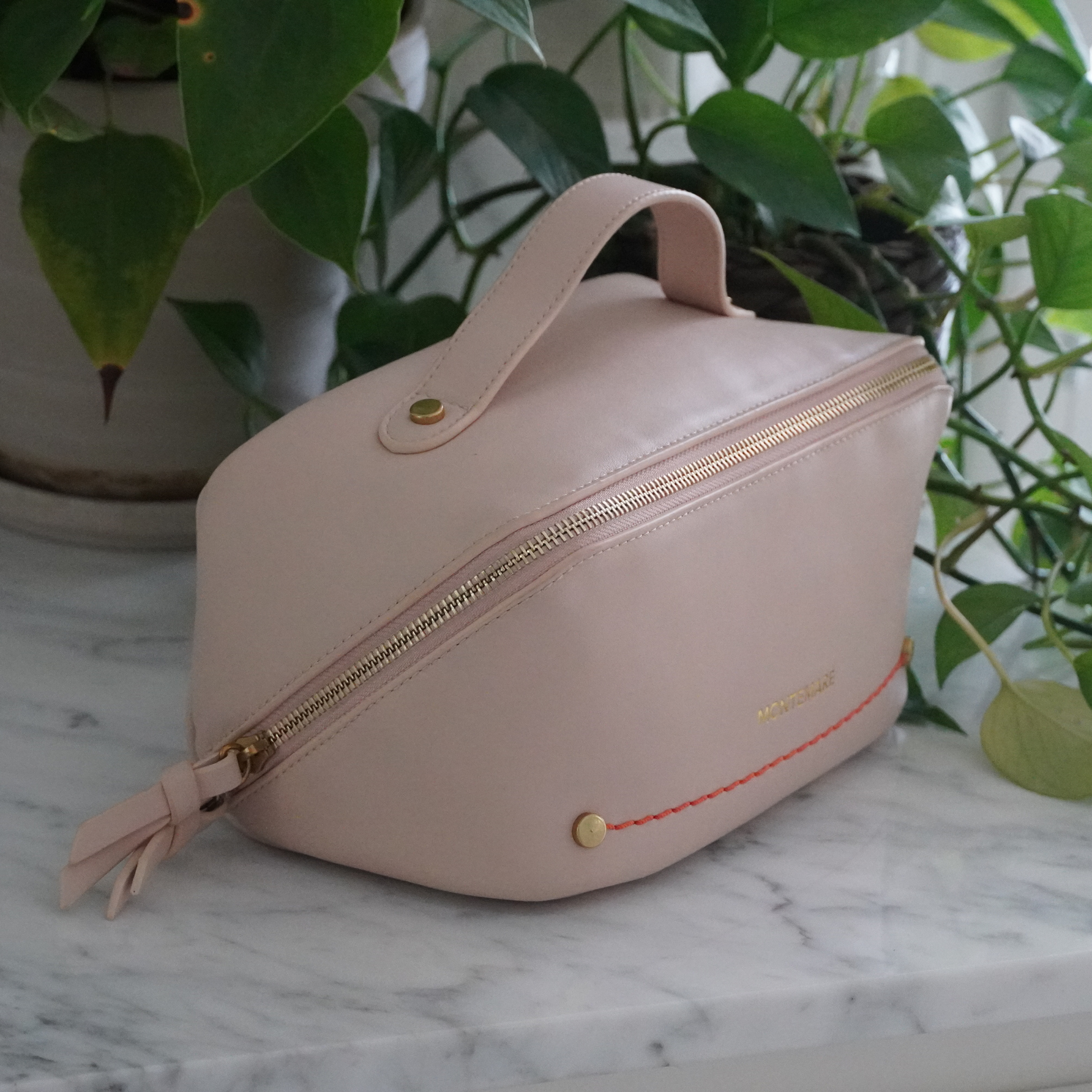 An ivory colored toiletry case with Montemare branding and an orange thread sitting on top of a marble surface with leafy background