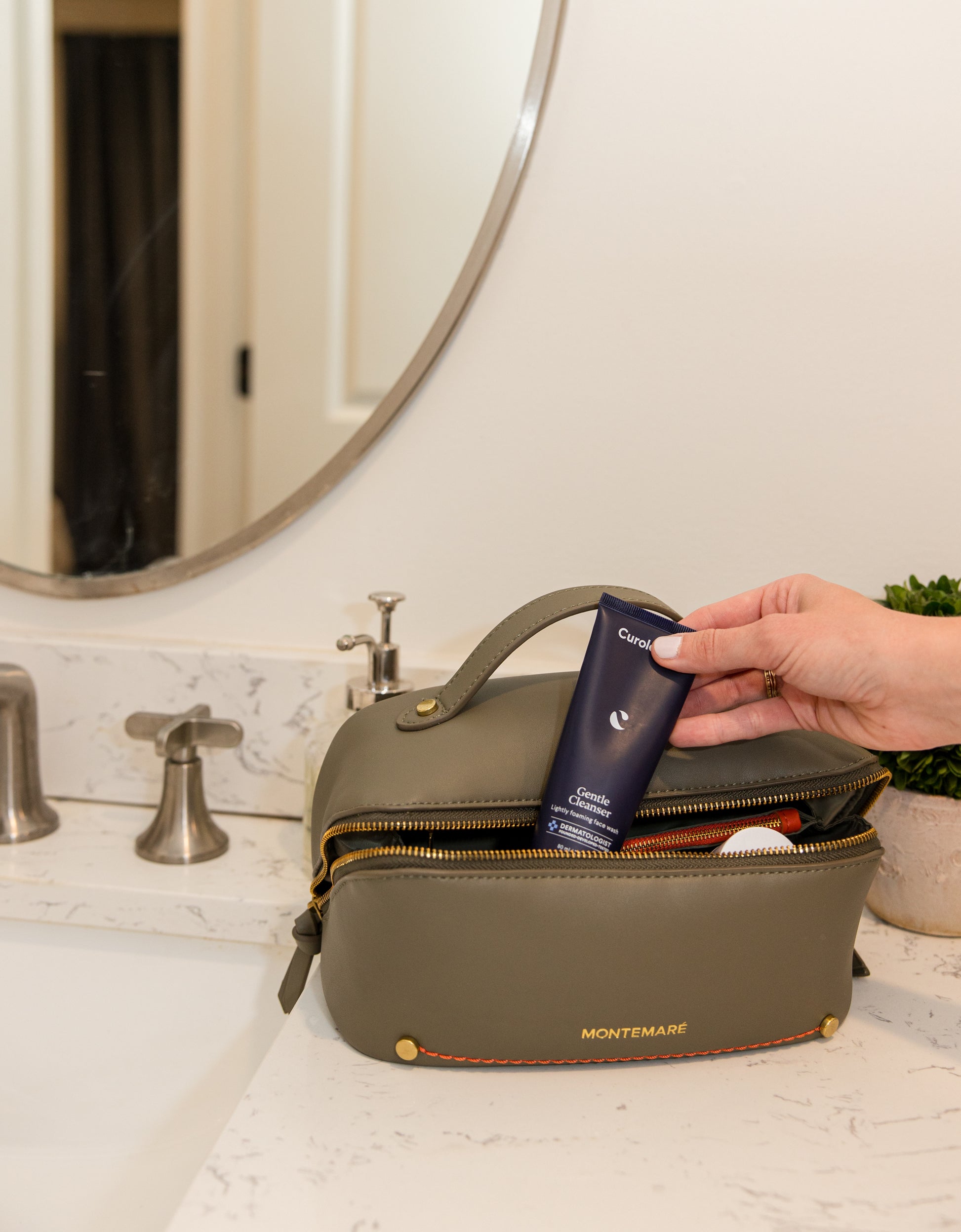 Person placing a tube of cream into a green toiletry bag on a bathroom counter.