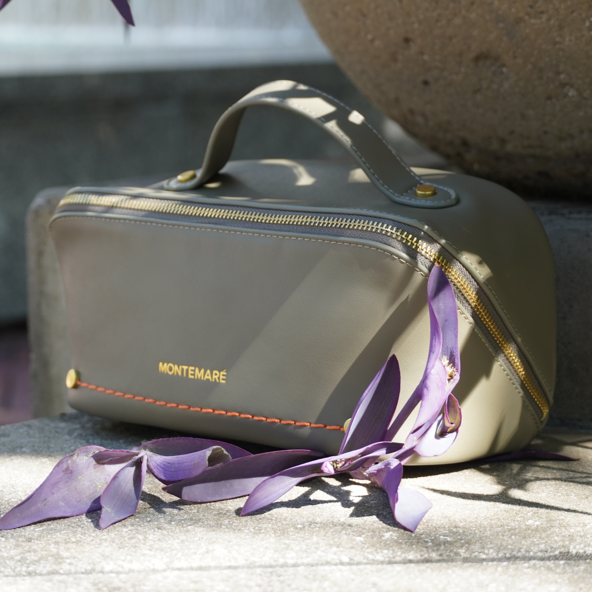 An olive green toiletry case with Montemare branding and orange thread on a stone background