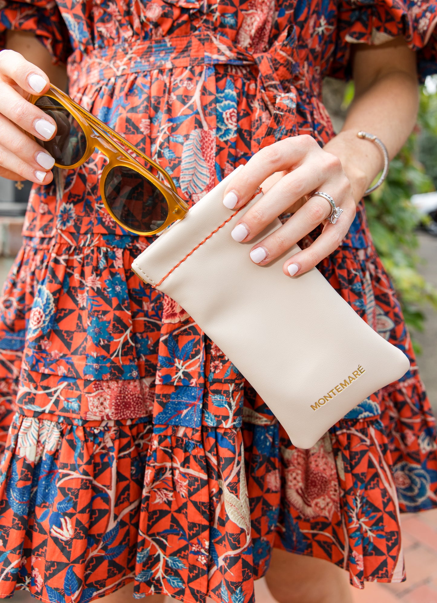 Person holding sunglasses and a beige pouch with 'Montemare' branding, wearing a colorful dress.