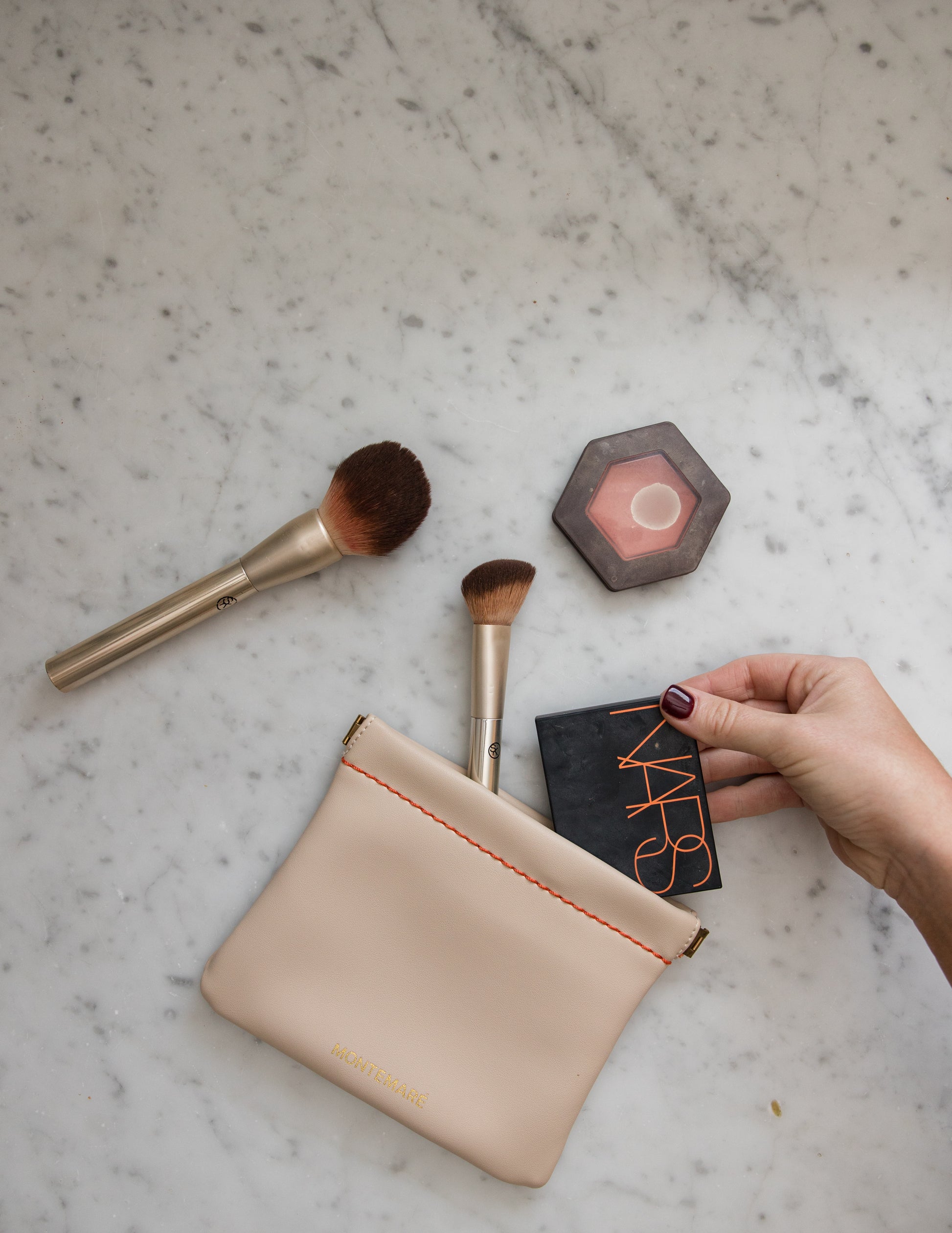 Makeup brushes and a white square pouch on a marble surface with a hand holding a small product.