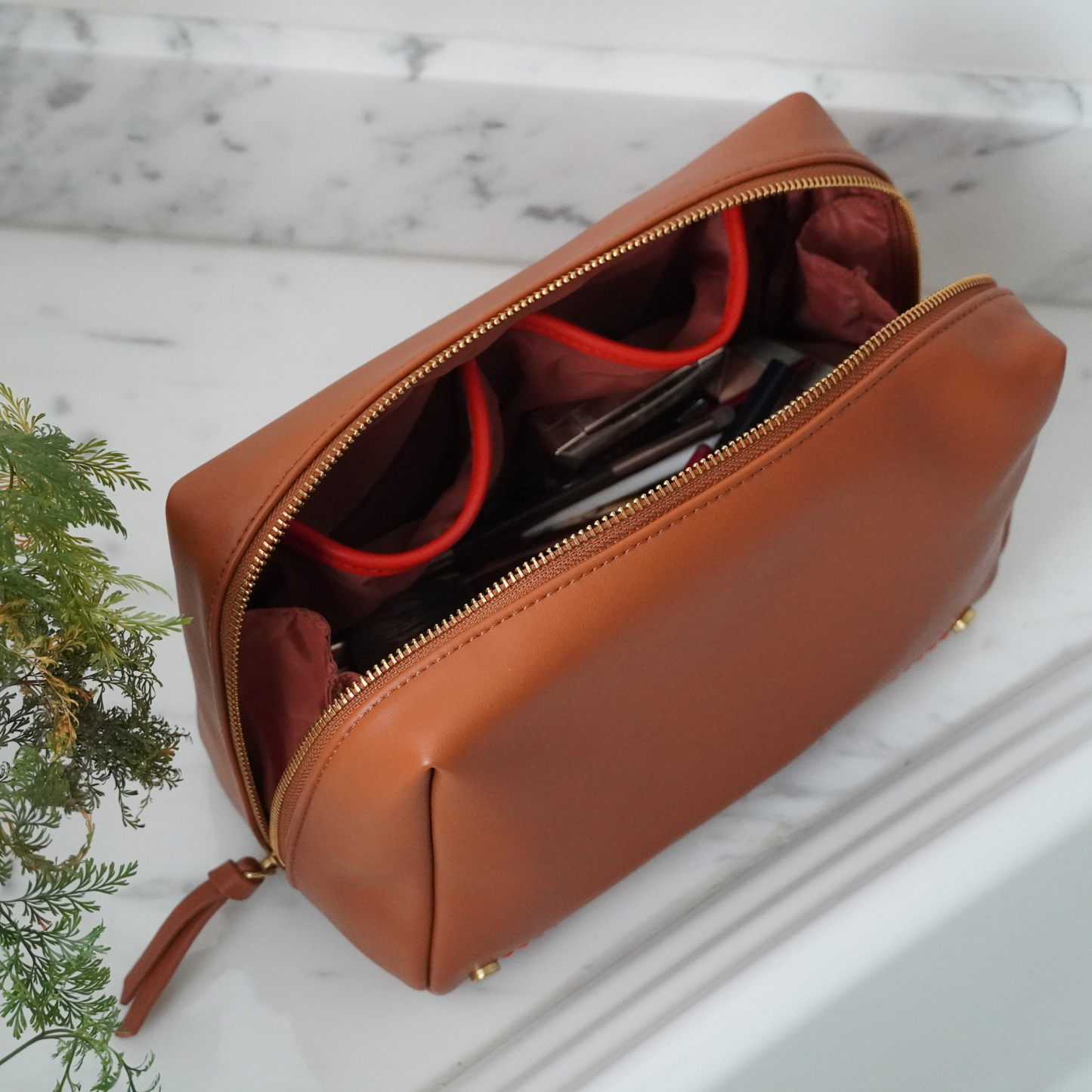 A brown Roma Standing Toiletry Case with a wide zipper opening, placed on a marble surface, with interior pockets visible and one zippered pocket closed.