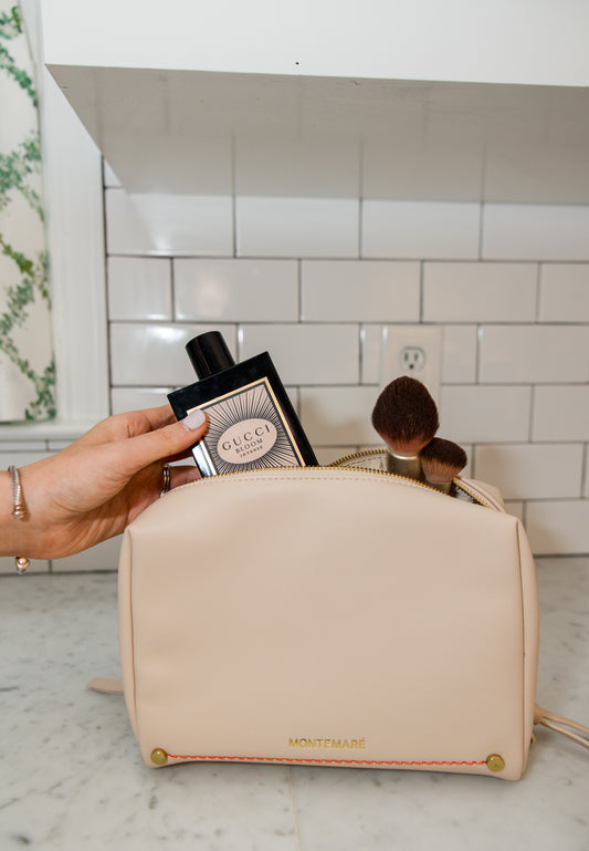 Person holding a Gucci perfume bottle next to a beige cosmetic bag with makeup items on a bathroom counter.