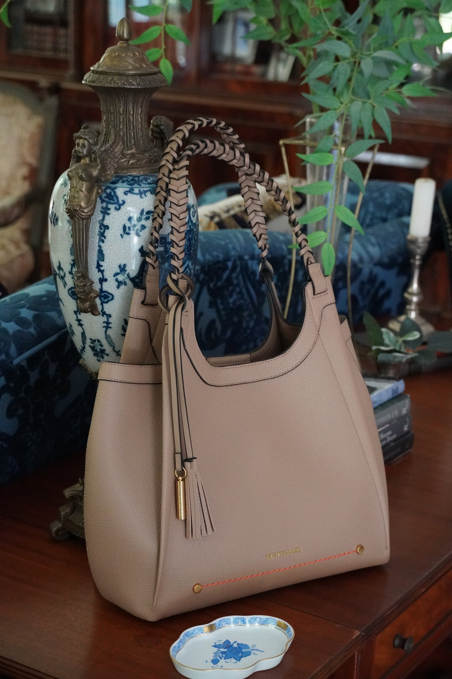 Beige handbag with tassels on a decorative surface with plants and books in the background