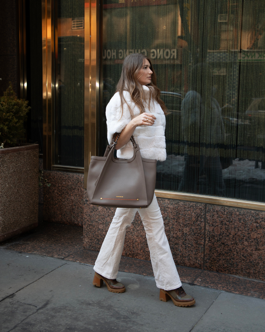 Woman in white puffer jacket and pants holding a brown tote bag, walking on a sidewalk.