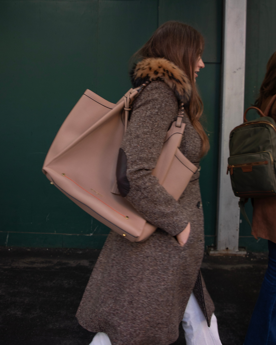 Woman in a long coat with a fur-lined hood carrying a beige tote bag against a green wall.