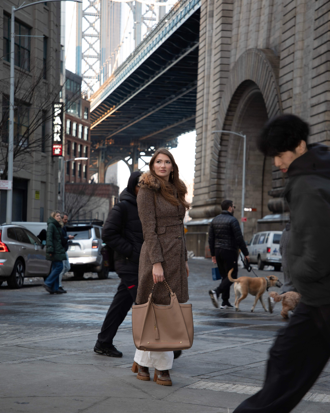 Woman with brown leather bag walking on a city street with a bridge in the background