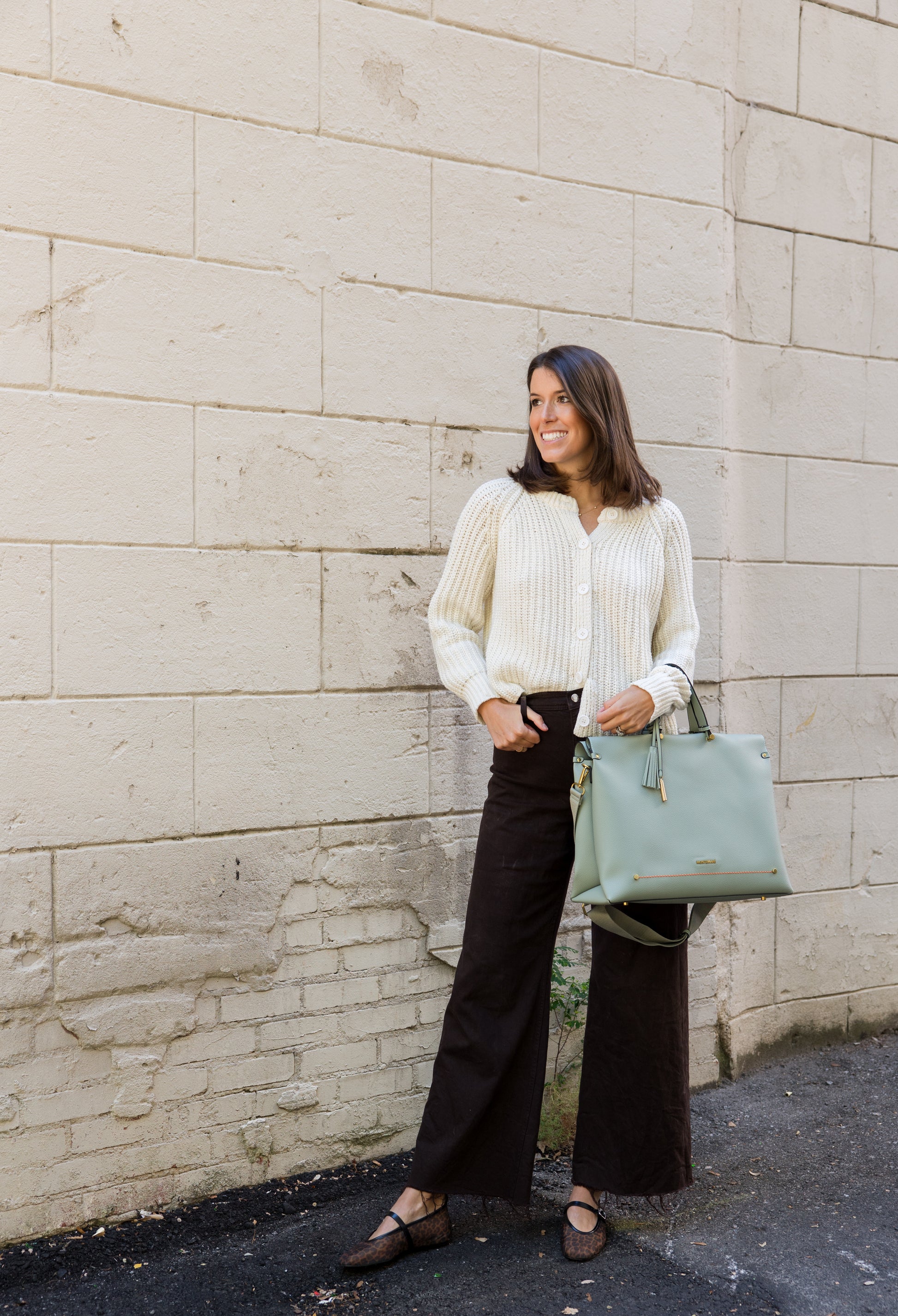 Woman in a white sweater and black pants holding a light blue handbag against a stone wall.