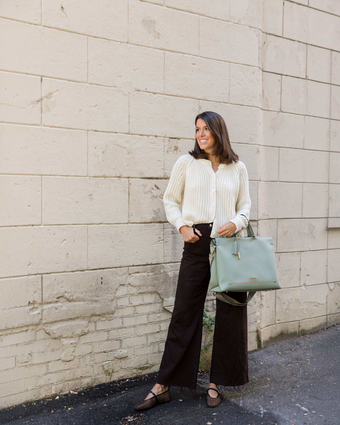 Woman in a white sweater and black pants holding a light blue handbag against a stone wall.