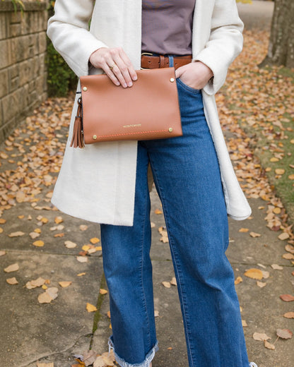 Person holding a brown clutch bag outdoors on a leafy ground
