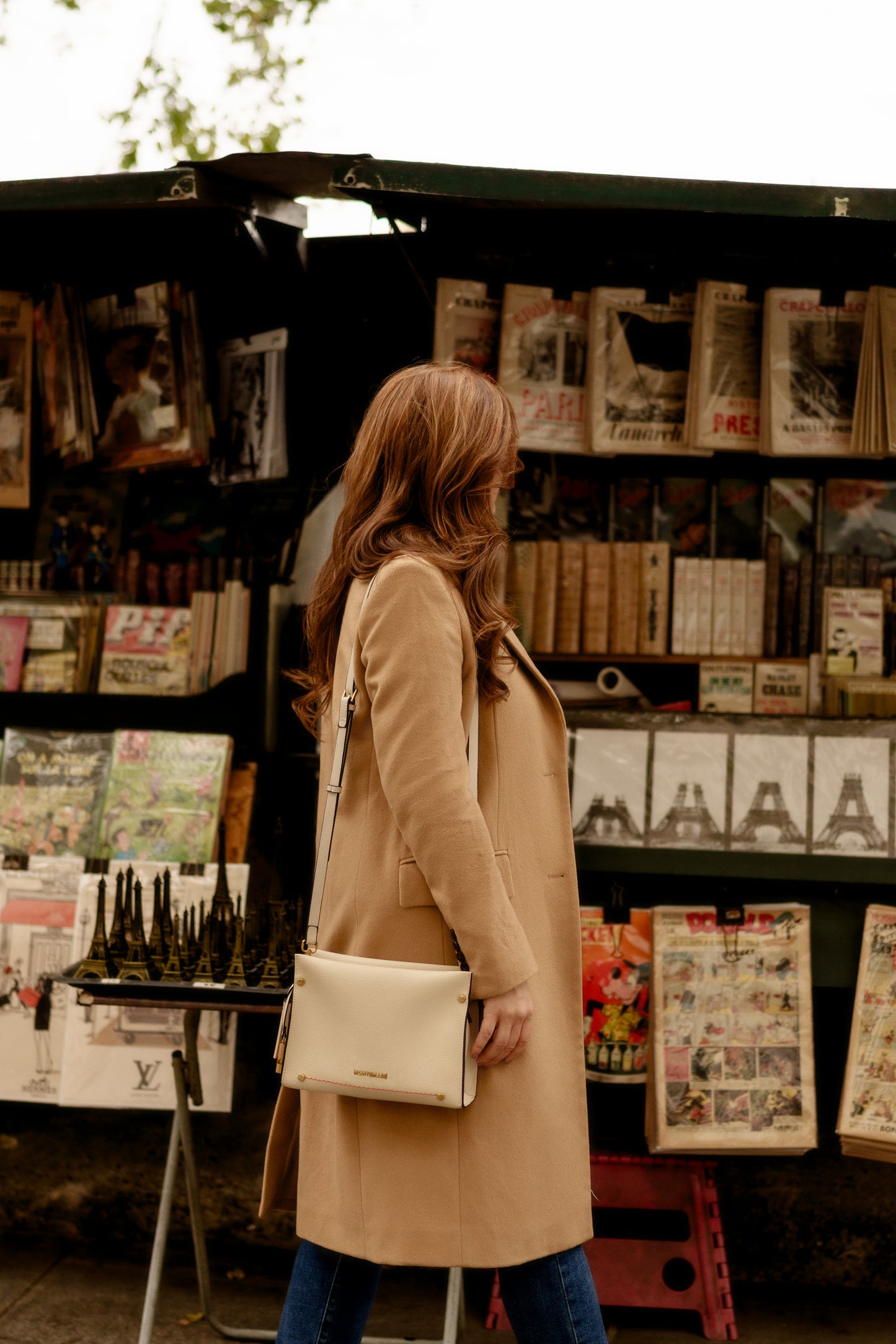 Woman in a beige coat with white handbag standing in front of a magazine stand with various publications.