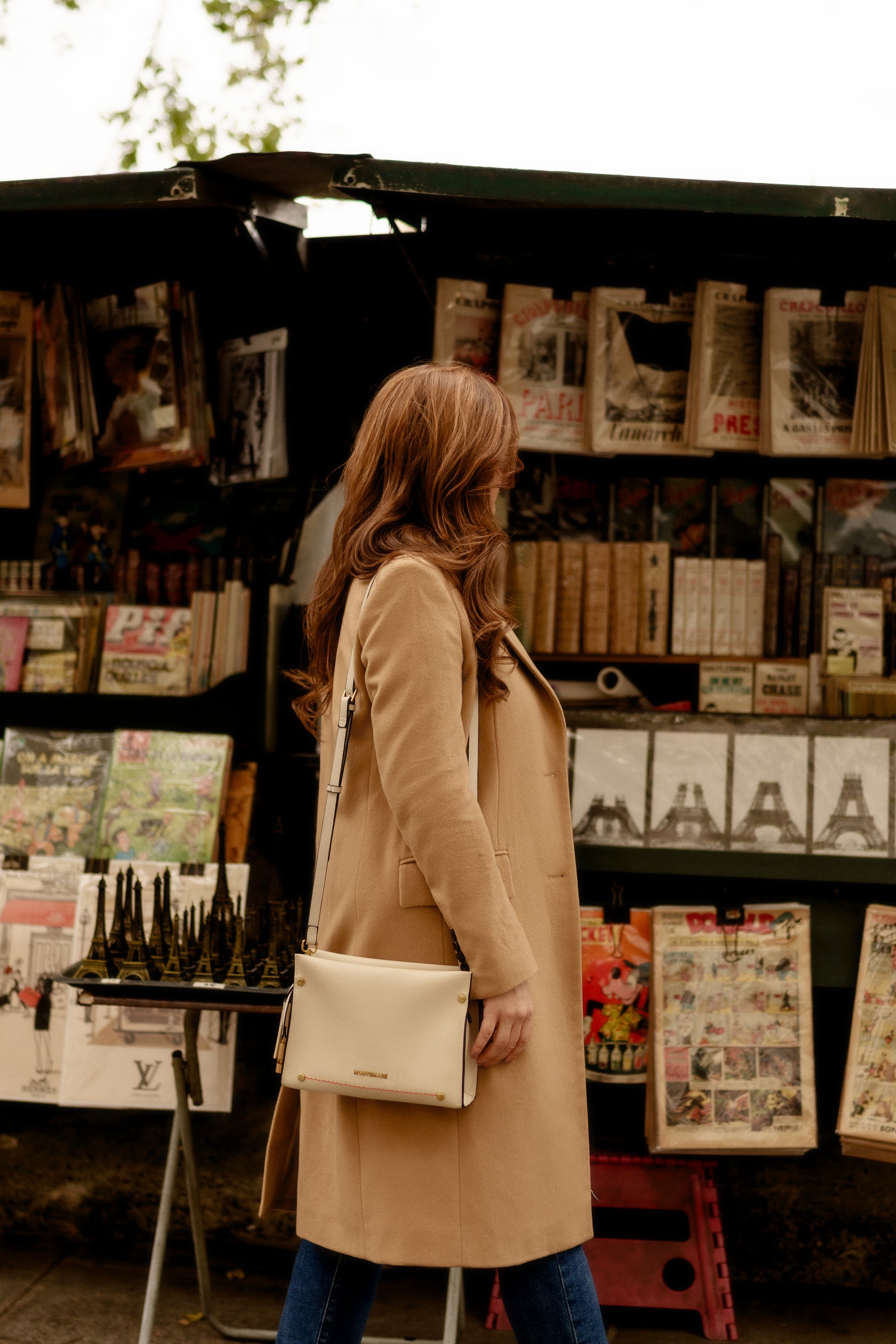 Woman in a beige coat with white handbag standing in front of a magazine stand with various publications.