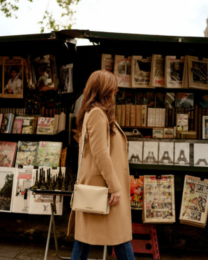 Woman in a beige coat with white handbag standing in front of a magazine stand with various publications.