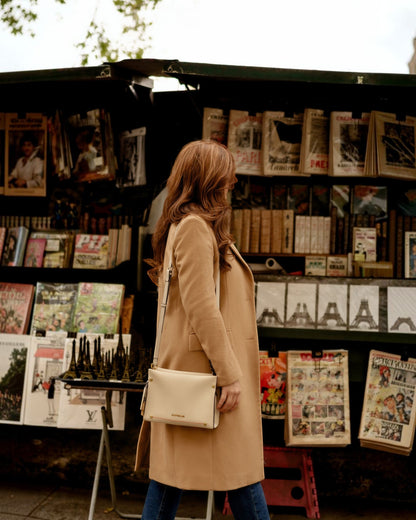 Woman in a beige coat with white handbag standing in front of a magazine stand with various publications.