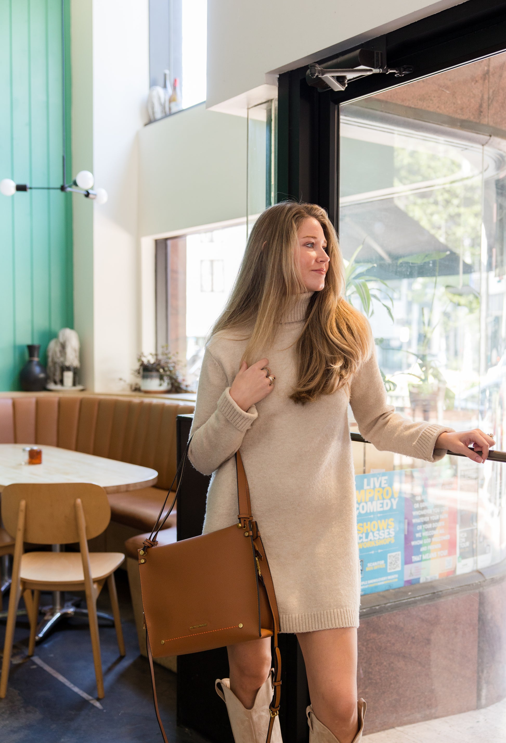 Woman with brown shoulder bag in a beige sweater dress standing in a modern cafe.