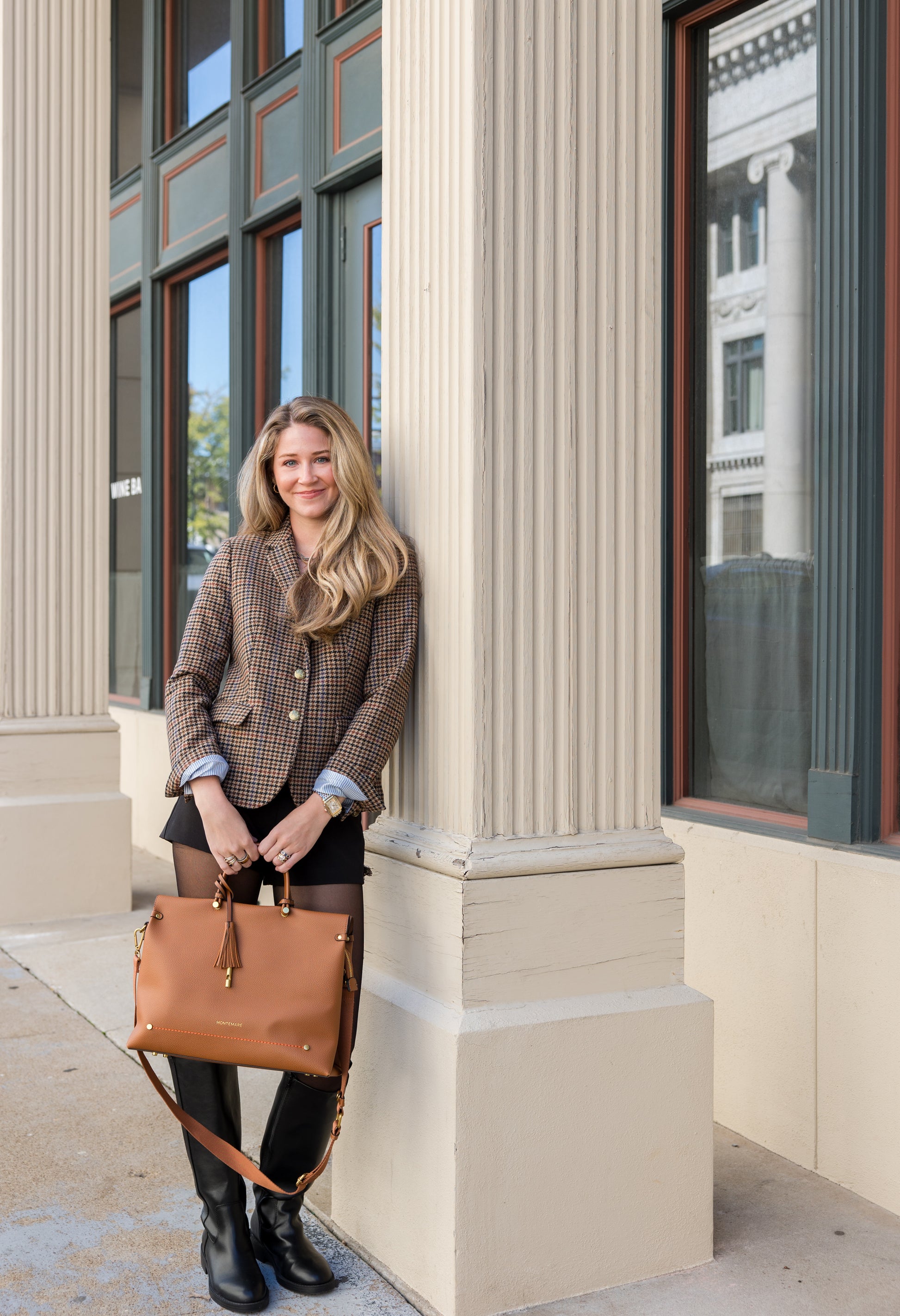 Woman holding a latte brown leather handbag standing against a building with columns.