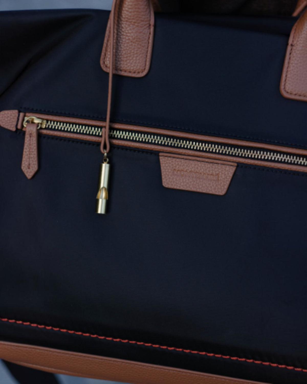 Close-up of a black Oslo Carry-On Bag with tan leather handles, zipper pulls, and trim, plus a small gold-toned zipper pull charm.