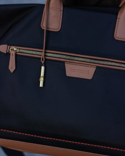 Close-up of a black Oslo Carry-On Bag with tan leather handles, zipper pulls, and trim, plus a small gold-toned zipper pull charm.