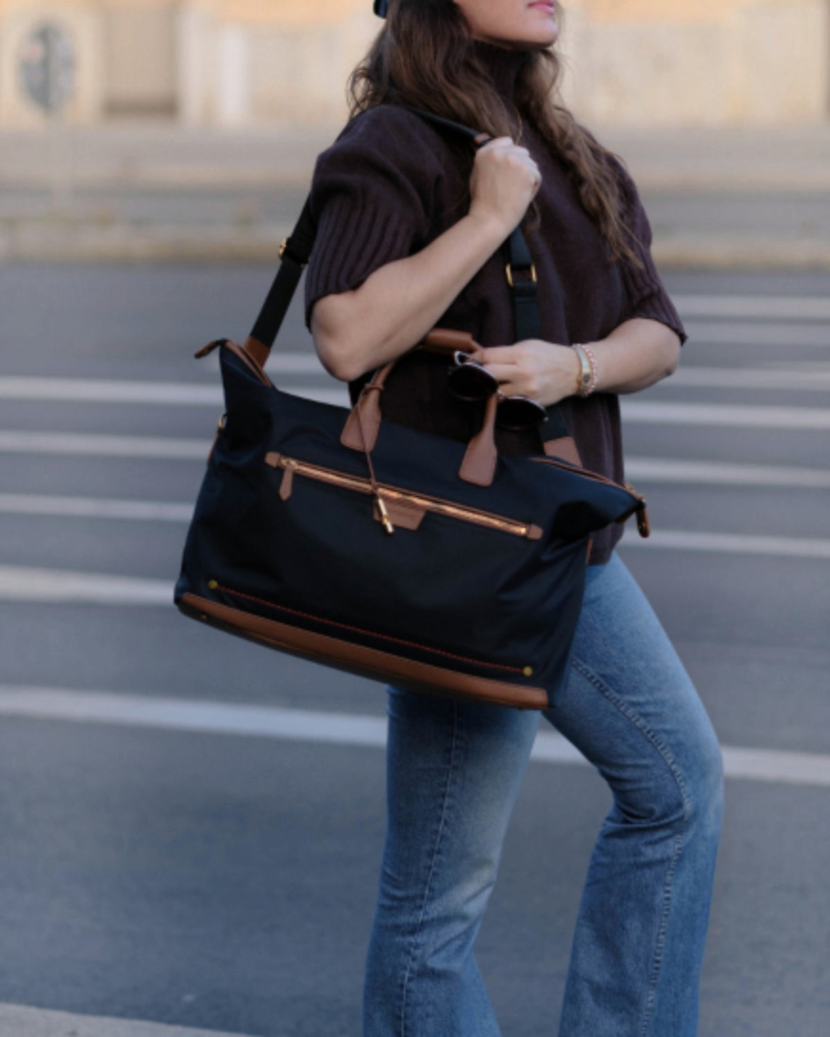A woman in a dark brown ribbed top and blue jeans carries a black Oslo Carry-On Bag with brown leather handles and trim, featuring a gold-tone front zipper, in an outdoor street setting.