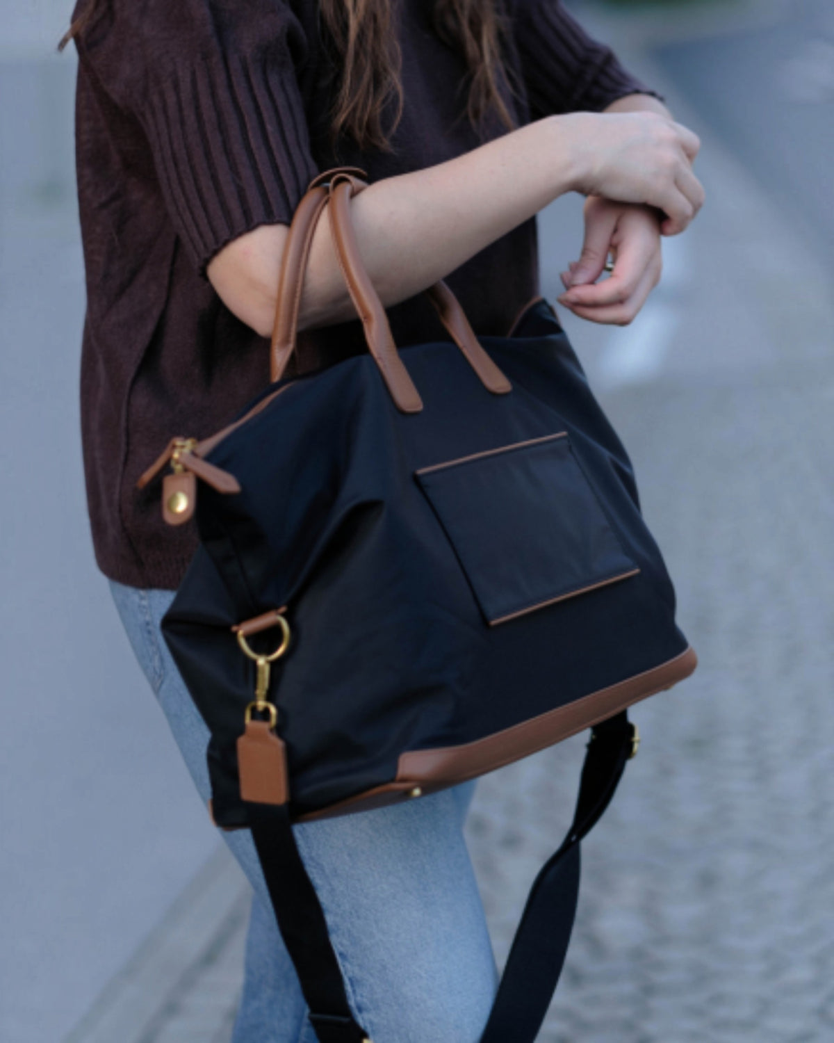 A person wearing a dark brown ribbed sweater and light blue jeans carries a black Oslo Carry-On Bag with brown leather handles, trim, and zipper pulls, shown outdoors on a paved street.