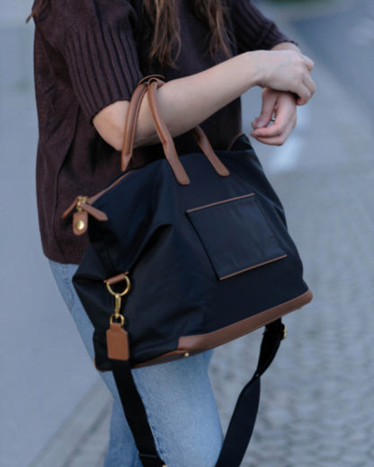 A person wearing a dark brown ribbed sweater and light blue jeans carries a black Oslo Carry-On Bag with brown leather handles, trim, and zipper pulls, shown outdoors on a paved street.
