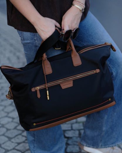 A person in blue jeans holds a black Oslo Carry-On Bag with brown leather handles and trim, featuring gold-tone hardware and a zippered front pocket, against a cobblestone street background.