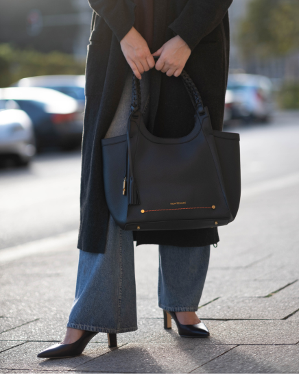 A person outdoors on a sidewalk carrying a Sedona Tote in Black with gold-tone hardware and braided top handles. The person is wearing a long dark coat, wide-leg blue jeans, and black heels.