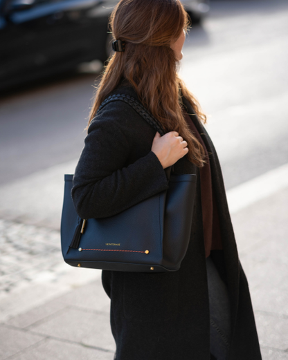 A woman in a black coat carrying a black Sedona Tote with gold hardware and red contrast stitching, photographed outdoors on a city street.