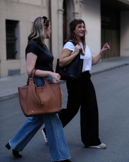 Two women walking on a city street, each carrying a Sedona handbag. The woman on the left wears a black t-shirt and blue jeans, while the woman on the right wears a white t-shirt, black wide-leg pants, and white sneakers.
