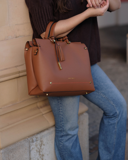 A woman wearing a dark brown sweater and blue jeans holds a Vienna Small Carry-On Tote in Cognac colorway with gold-tone hardware and a tassel detail against a beige stone wall.
