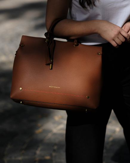 A person carrying a Vienna Small Carry-On Tote in Cognac colorway outdoors. The bag has gold-tone hardware, a top handle, and a visible 'Montemare' logo on the front.